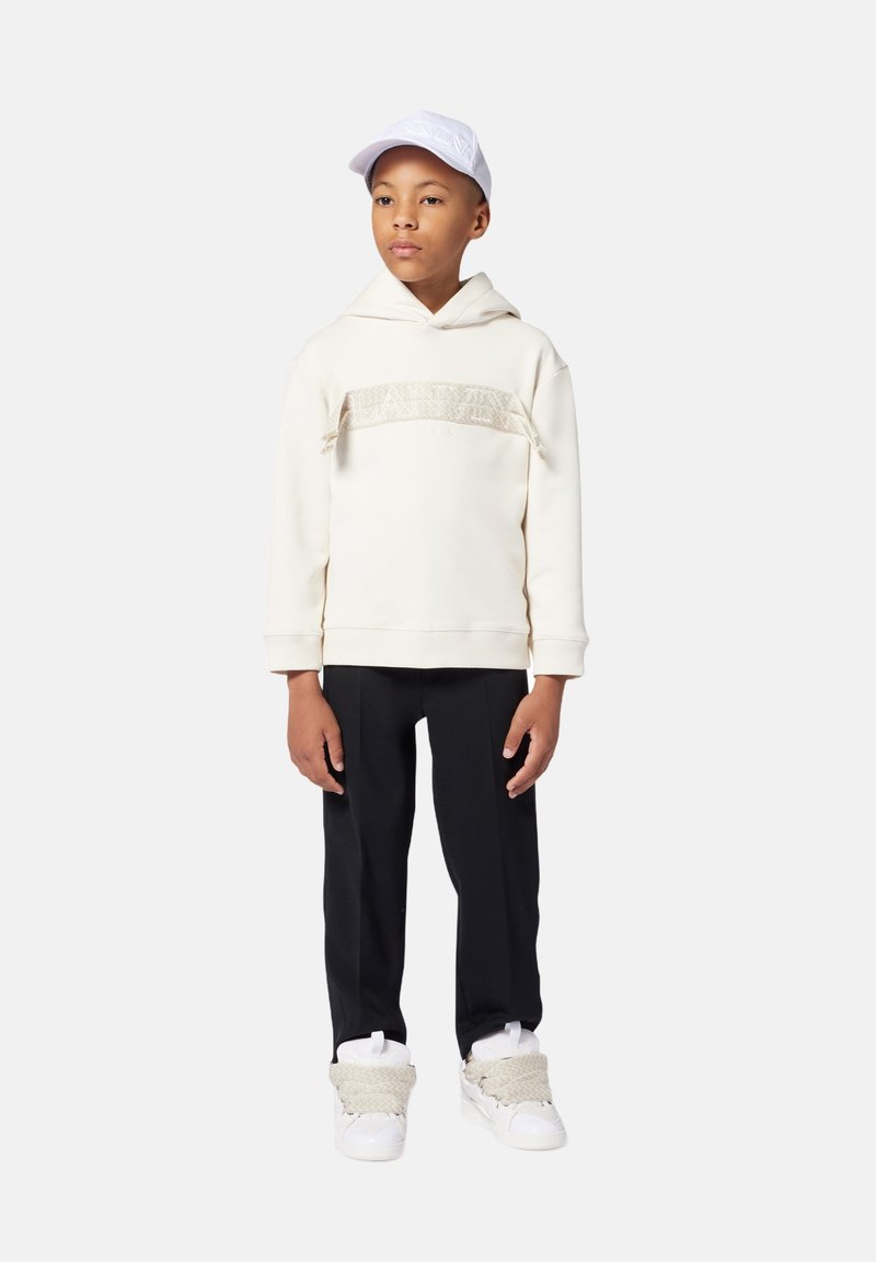 Felpa crema con banda texturizzata, pantaloni neri e sneakers bianche con dettagli intrecciati. Il bambino indossa un cappellino bianco. Outfit semplice e informale.