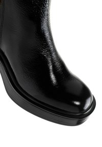 Gros plan sur l'avant d'une botte en cuir noir brillant à la forme arrondie et à la semelle épaisse en caoutchouc sur fond blanc.