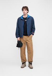 Ναυτικό σακάκι με μπλε κολάρο από φλις, μαύρο μπλουζάκι με λογότυπο, χακί παντελόνια και γκρι μπότες. Κρατάει μια μικρή μαύρη τσάντα.