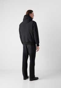 Μαύρο μπουφάν με κουκούλα, ραβδωτές μανσέτες και στρίφωμα, συνδυασμένο με σκούρες γκρι τζιν με ίσια γραμμή και μαύρα παπούτσια. Πίσω άποψη του ντυσίματος.