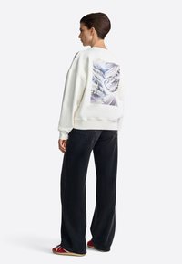 Felpa bianca con grafica paesaggistica sulla schiena, abbinata a jeans neri a gamba larga e scarpe rosse. Materiale morbido, vestibilità comoda.