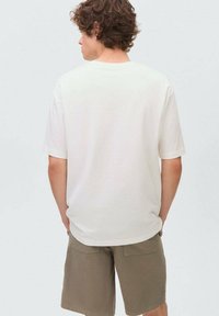 Bílý tričko s krátkým rukávem a volným střihem. Hladká bavlněná tkanina, lehce prodloužený zadní díl. Nosené s olivově zelenými šortkami.