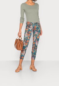 Haut à manches longues rayé vert, pantalons courts à imprimé floral, sandales à lanières beiges et un sac à main marron. Couleurs vives et motifs détaillés.