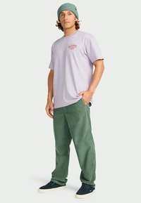 Man wearing light purple graphic t-shirt, green pants, teal beanie, and black sneakers, standing with one hand in pocket against plain background.