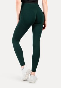 Ciemnozielone legginsy z elastycznego materiału, z wysokim stanem i gładką fakturą. Połączone z białymi butami sportowymi.