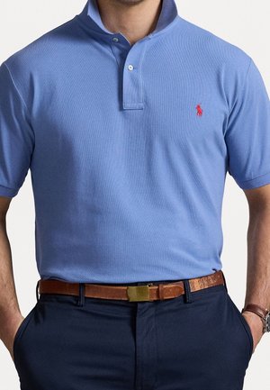 Mand iført en lyseblå polo med et lille rødt logo, marineblå bukser og et brunt læderbælte med en guldlås.