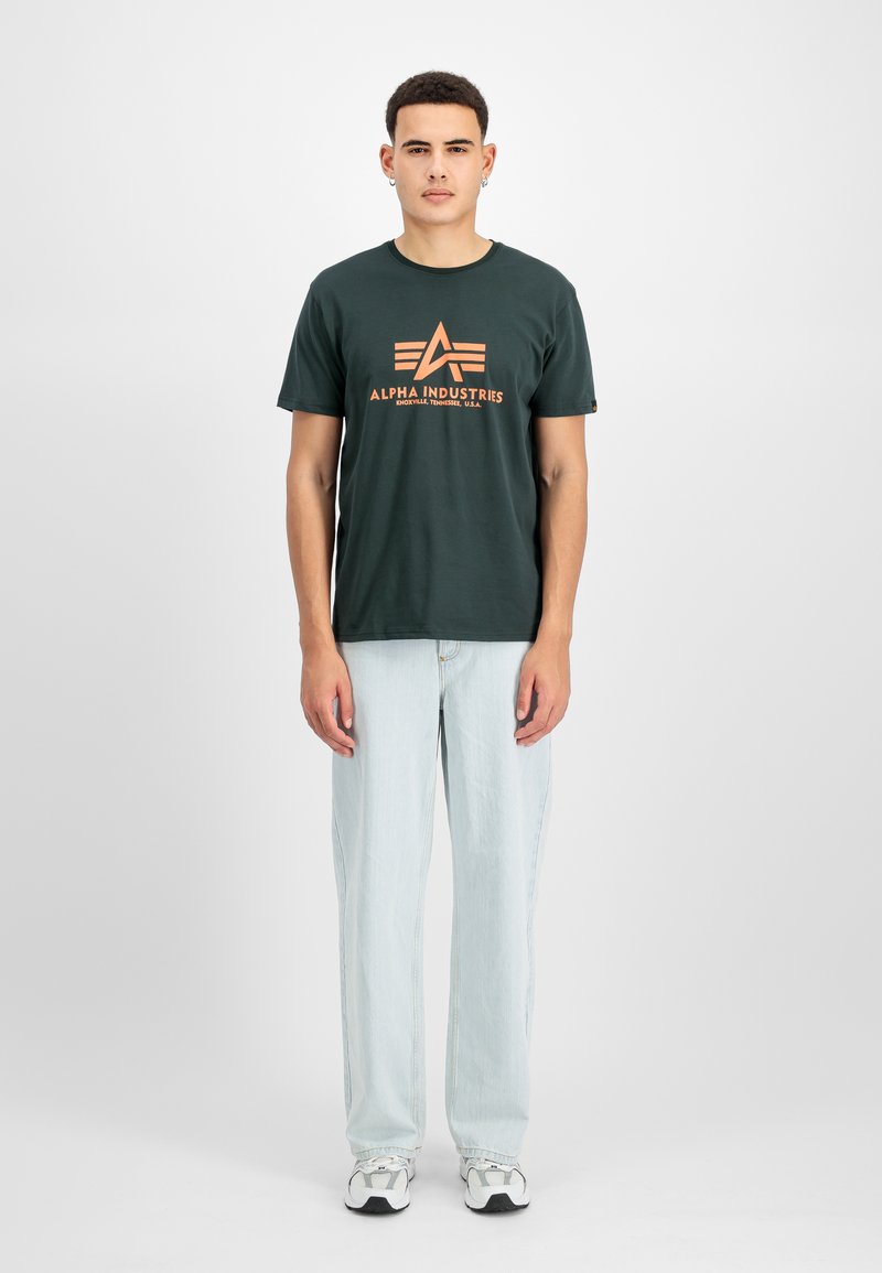 Junger Mann, der nach vorne blickt, trägt ein dunkelgrünes Alpha Industries T-Shirt, hellblaue Jeans und weiße Sneaker vor einfachem Hintergrund.