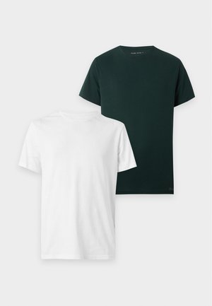 Homme portant une chemise à manches courtes vert foncé, un jean bleu clair et des baskets marron, debout avec une main dans la poche sur fond blanc.