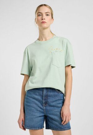 Lichtgroen katoenen t-shirt met een borstzak, voorzien van gele bloemenborduursels. Gecombineerd met blauwe denim shorts. Ontspannen pasvorm.
