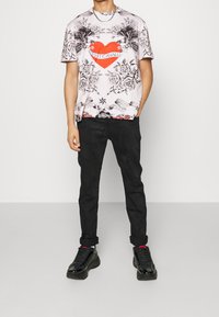 Man som bär en vit grafisk T-shirt med svarta blommönster och örndesigner samt ett rött hjärta, svarta byxor och svarta sneakers med röda strumpor.