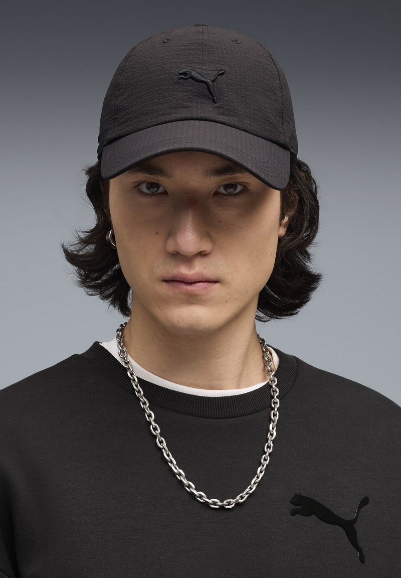 Jeune personne aux cheveux mi-longs foncés, portant une casquette noire Puma, un pull noir avec le logo Puma, une chaîne en argent et une boucle d'oreille.