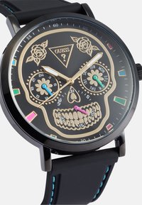 Relógio analógico preto com um design de caveira, acabamentos em dourado, detalhes florais e marcadores de horas coloridos. Bracelete de borracha com costura azul.