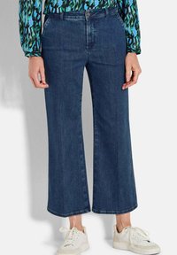 Pantalon en denim bleu foncé à jambe large avec une taille mi-haute et une silhouette droite, associé à des baskets blanches sur un fond uni.