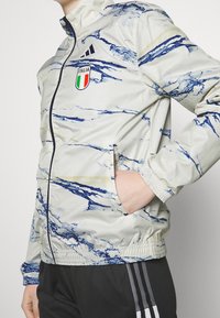 Lätt, mönstrad jacka i ljusgrått med blå och gul marmor-design; har dragkedja fram, krage och Italiens flagga på bröstet.