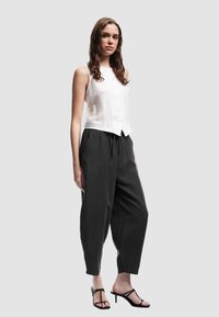 Jonge vrouw met lang bruin haar, gekleed in een mouwloze witte blouse, losse zwarte cropped broek en zwarte gespiseerde pumps.