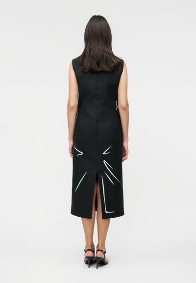 Γυναίκα όρθια, φοράει μαύρο midi φόρεμα χωρίς μανίκια με λευκές αφηρημένες γραμμές και σχίσιμο στην πλάτη, συνδυασμένο με μαύρα πέδιλα με λουράκια.