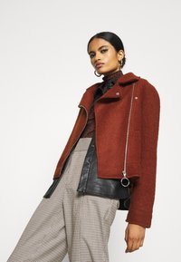 Giacca corta in misto lana marrone con un davanti in pelle nera a contrasto, dotata di chiusura con zip e colletto rialzato. Top con motivo e pantaloni a quadri.