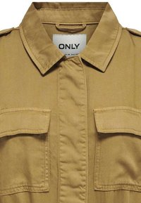 Chemise beige à manches courtes avec col, présentant deux poches avant, en tissu lisse avec un boutonnage sur le devant et des coutures renforcées.