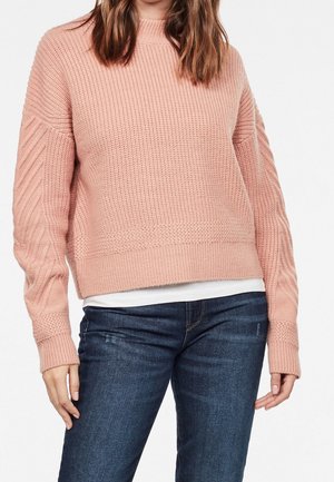 Ljusrosa stickad tröja med ribbat mönster, rund hals och flätstickade ärmar. Matchad med mörkblå jeans.