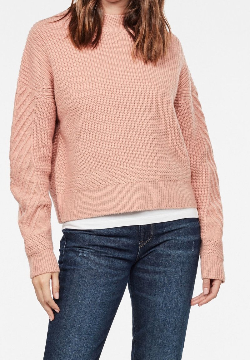 Ljusrosa stickad tröja med ribbat mönster, rund hals och flätstickade ärmar. Matchad med mörkblå jeans.