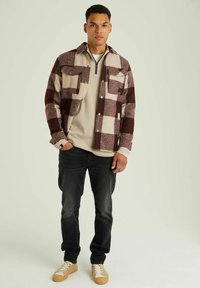 Geruite flanellen overhemd in bordeauxrood en crème, gedragen over een lichtbeige lange mouwen shirt, gecombineerd met donkere jeans en beige sneakers.