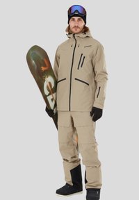 Giacca da neve beige con dettagli neri, pantaloni beige coordinati e guanti neri. Tiene uno snowboard con un design grafico. Il portatore indossa occhiali protettivi.