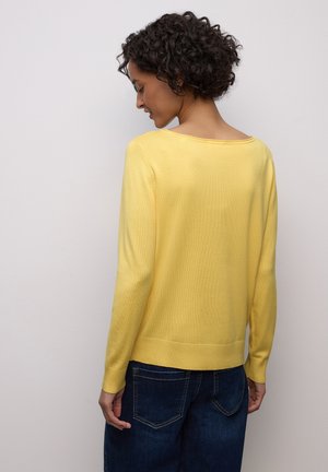 Pull jaune en maille avec un large col rond et des manches longues. Comprend des poignets et un ourlet côtelés. Porté avec un jean bleu foncé.