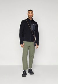 Black fleece jacket with a zippered pocket, olive green pants, black sneakers, and a small black pouch with a shoulder strap.