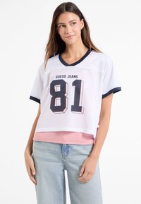 Hvid mesh crop top med marineblå detaljer, prydet med et iøjnefaldende "81" grafik og "GUESS JEANS" tekst. Burd over en lyserød lagdelt skjorte. Casual pasform.