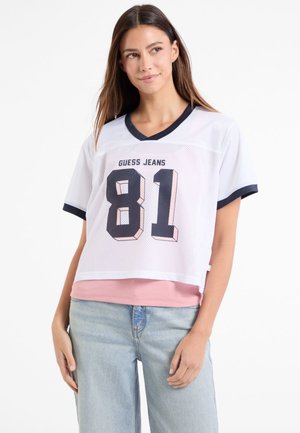 Biely priehľadný crop top s námorníckymi akcentmi, s odvážnou grafikou "81" a textom "GUESS JEANS". Nosené cez ružovú vrstvenú košeľu. Casual strih.