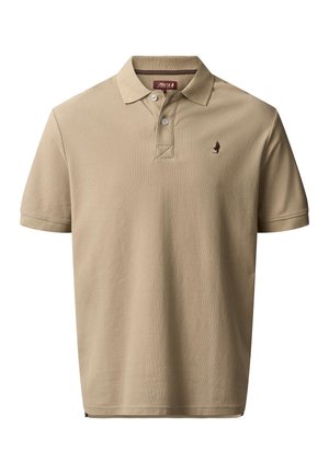 Homme aux cheveux mi-longs et barbe, portant un polo beige et un pantalon bleu marine, mains dans les poches, regardant sur le côté, avec des bras tatoués.