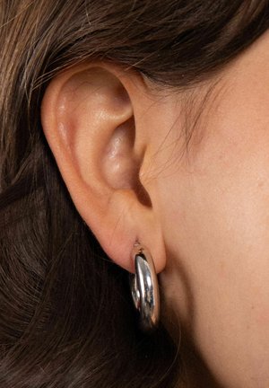 Nahaufnahme eines Ohres einer Person mit langem braunem Haar, die einen dicken, glänzenden silbernen Creolenohrring am Ohrläppchen trägt.