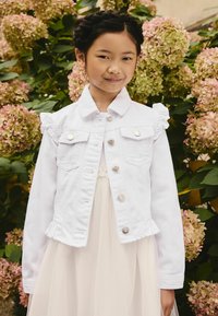 Nuori tyttö valkoisessa denimitakissa ja kermaisessa mekossa seisoo pinkkien ja vihreiden hortensioiden edessä, hymyillen lempeästi.