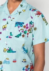Camicia azzurra a maniche corte con una varietà di motivi colorati che includono insetti, fiori e simboli del sole, dotata di taschino sul petto.