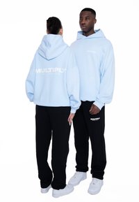 Sudaderas azul claro con impresión de logotipo blanco, combinadas con pantalones de chándal negros. La sudadera tiene un corte amplio y un gran bolsillo frontal.