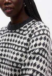 Stickad tröja i svart och vitt med geometriska mönster; har en rund halsringning och texturerat tyg. Närbild på ärmdetalj.
