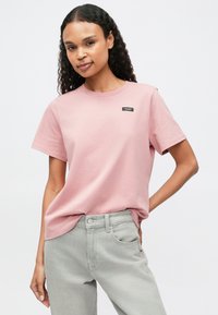 Φωτεινό ροζ κοντομάνικο μπλουζάκι από βαμβάκι, με στρογγυλή λαιμόκοψη και μικρή μαύρη λογότυπο ραφή στο στήθος. Συνδυάζεται με γκρίζα τζιν.