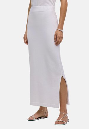 Vrouw die een witte enkellange rok met een zijsplit en witte platte sandalen met bandjes draagt, staand tegen een effen achtergrond.