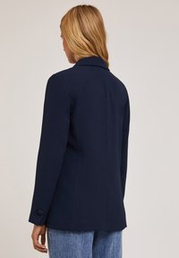 Granatowy blezer o dopasowanym kroju, z klapami, pojedynczym guzikiem na mankietach i gładkiego materiału, zestawiony z jasnymi dżinsami.