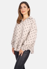 Light beige button-up shirt featuring a textured fabric with black heart patterns, a collar, and loose sleeves. Worn with dark pants.