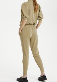 Khaki loungewear set obsahující top s nabíranými rukávy a zúžené kalhoty, vyrobený z měkké látky s bočními kapsami. Doplňuje ho černé nazouvací boty.