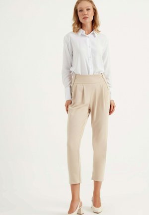 Weißes Hemd mit Knöpfen, kombiniert mit beigen, plissierten Hosen mit seitlichem Reißverschluss, glatter Textur und cropped Länge. Klassische Absatzschuhe runden den Look ab.