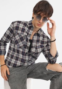 Camicia a scacchi nei colori grigio, blu navy e rosso; due tasche frontali, maniche lunghe. Indossata con jeans grigi strappati e occhiali da sole rotondi.
