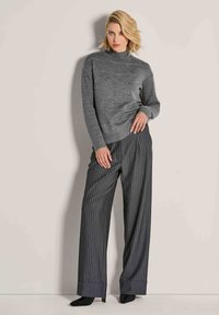 Frau mit kurzem blonden Haar, die einen grauen Rollkragenpullover und dunkelgraue gestreifte weite Hosen trägt, sowie schwarze spitze Schuhe.