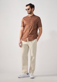 Short-sleeve brown shirt with vertical white stripes, buttoned, paired with cream trousers and white sneakers. Man wearing sunglasses.