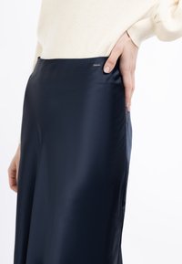 Jupe en satin bleu marine avec une texture lisse, une fente latérale et une étiquette de marque près de la ceinture. Le tissu a une finition brillante.