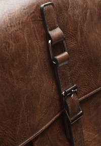 Borsa a tracolla in pelle marrone con superficie texturizzata, dotata di una fibbia in metallo e di una cinghia per la chiusura. Primo piano delle parti metalliche e dei dettagli.