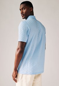 Světle modré polo tričko s krátkým rukávem, s texturovaným diamantovým vzorem, s límečkem a volným střihem, v kombinaci s světlými kalhotami.