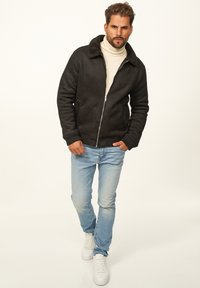 Schwarze Jacke aus Schaffellimit mit Reißverschluss, kombiniert mit hellblauen Jeans und weißen Turnschuhen. Die Jacke verfügt über einen breiten Kragen und gerippte Bündchen.