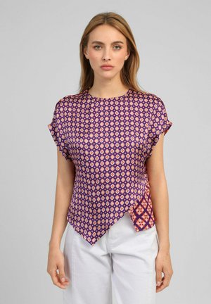 Femme portant un chemisier à manches courtes violet à motif floral avec un ourlet asymétrique et un pantalon blanc, debout devant un fond gris uni.